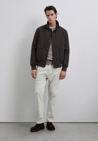 Joven con una chaqueta marrón oscuro, un suéter beige claro, pantalones blancos y botas marrón oscuro, de pie contra un fondo gris claro.