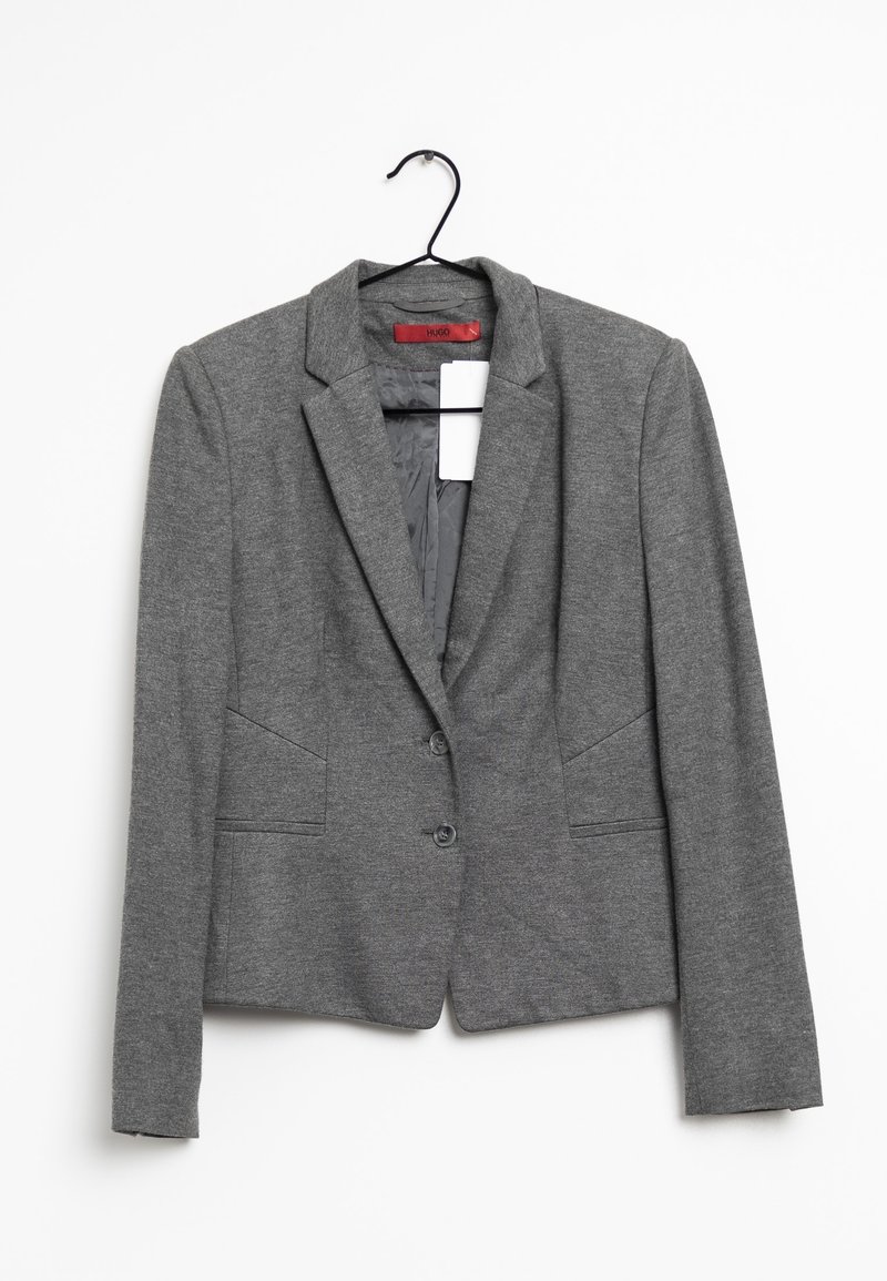 Blazer gris sur mesure avec deux boutons à l'avant et poches latérales, suspendu à un cintre noir sur fond blanc.