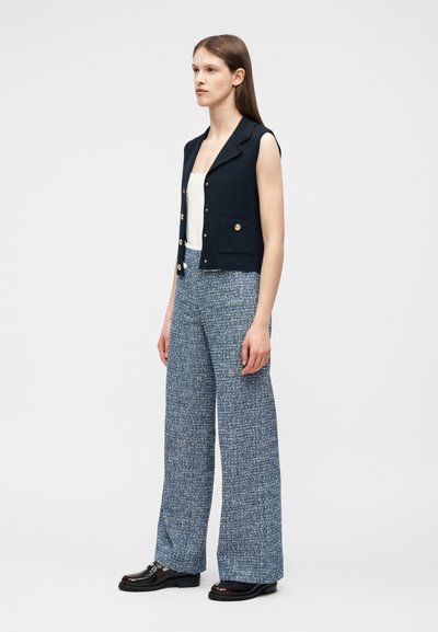 Γυναίκα όρθια με αμάνικο μπλε σκούρο γιλέκο με χρυσά κουμπιά, λευκό τοπ, μπλε παντελόνι από υφαντό ύφασμα με φαρδιά πόδια και μαύρα μοκασίνια σε λευκό φόντο.