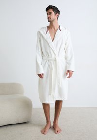 Accappatoio bianco in spugna con cappuccio, fascia interna a contrasto, cintura annodata, che arriva a metà polpaccio e maniche lunghe.