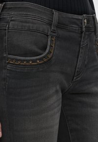 Μαύρο τζιν από denim με μπροστινές τσέπες διακοσμημένες με μικρά στρογγυλά ορειχάλκινα καρφιά και αντίθετες καφέ ραφές, φορεμένο με μαύρο ριμπ πουκάμισο.