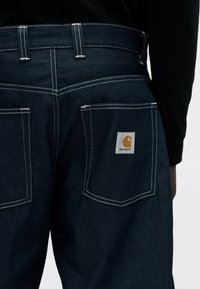 Πλάγια όψη σκούρου μπλε τζιν παντελονιού με λευκή ραφή και τετράγωνο λογότυπο Carhartt στο δεξί πίσω τσεπάκι, φορεμένο με μαύρο μακρυμάνικο τοπ.