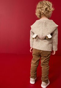 Beige gebreide trui met een puntige capuchon en konijnenoren accenten, gecombineerd met een brown broek en lichte schoenen, gezien vanuit de achterkant.