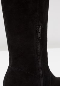 Yhden jalanmukaisista mustista mokkapohjaisista ylipolvista saappaista, jotka sisältävät sivuvetoketjun ja mukavan pinnan, jossa on hienovaraisia ompelusaumoja.