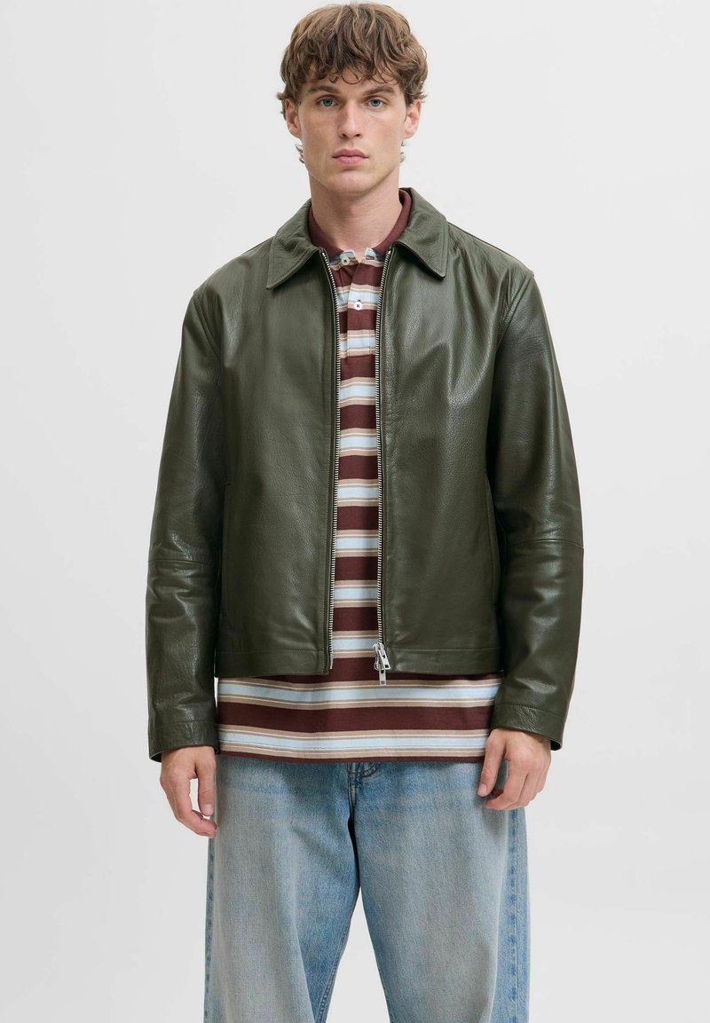 Chaqueta de cuero verde oliva con cremallera frontal, cuello y mangas largas. Llevada sobre una camisa a rayas en tonos marrón y beige, combinada con jeans azules.