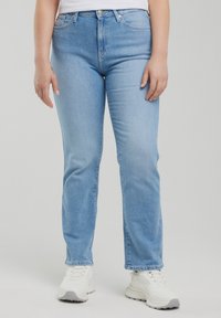 Persona, kas valkā gaiši zilas taisnu kāju džinsas, baltas sporta kurpes un baltu kreklu, stāvot pret vienkāršu fonu.