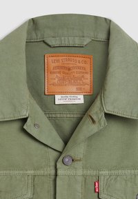 Chaqueta Levi's verde oliva con cuello de botón, cierre de metal a presión y una etiqueta de cuero marrón con el logotipo y detalles de cuidado.