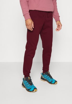 Persona che indossa pantaloni della tuta borgogna, una felpa rosa polveroso e scarpe sportive blu, nere e gialle, in piedi su un pavimento chiaro e semplice.