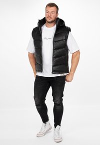 Zwarte puffervest met een capuchon, rits aan de voorkant en zijzakken, gedragen over een wit t-shirt en gecombineerd met zwarte gescheurde spijkerbroeken en witte sneakers.