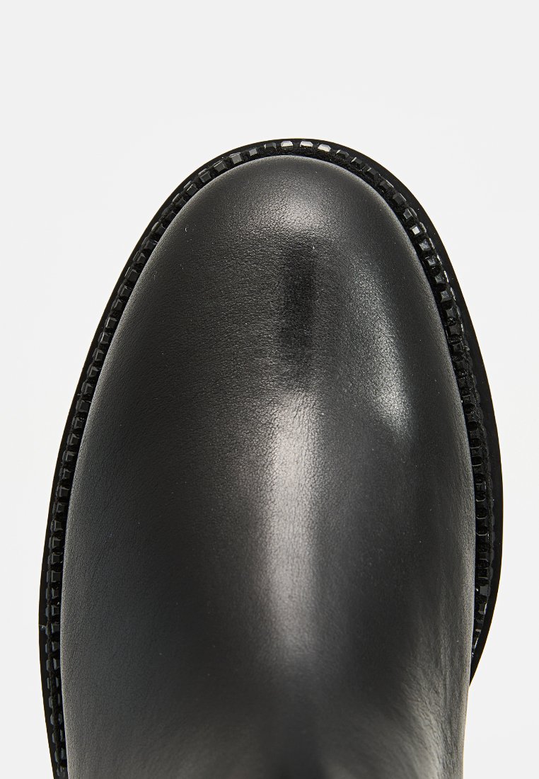 Bota de cuero negro con textura lisa, puntera redondeada y costuras en negro contrastante a lo largo del borde. Sin herrajes ni patrones visibles.