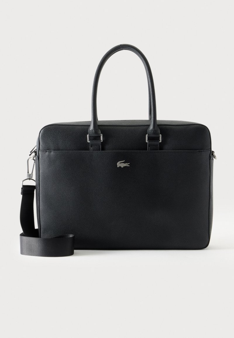 Mallette en cuir noir avec deux poignées, bandoulière amovible et petit logo crocodile argenté sur la poche avant.