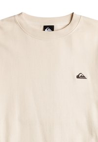 Beige mikina vyrobená z mäkkého bavlneného materiálu, má rebrovaný okrúhly výstrih a malé tmavé logo umiestnené na ľavej hrudi. Hladká textúra.