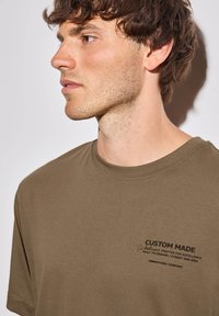 Olivově zelené bavlněné tričko s kulatým výstřihem a krátkým rukávem. Na hrudi je černý textový grafický prvek, který uvádí značku a vlastnosti pohodlí.