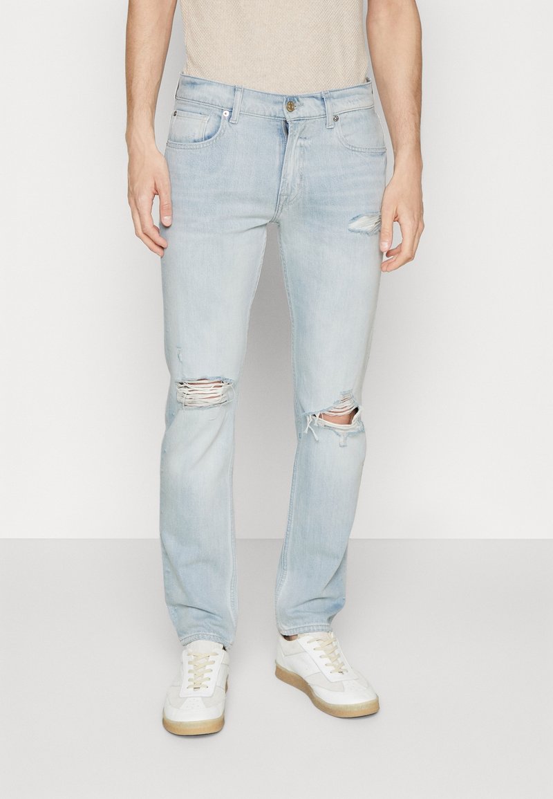 Vaqueros azul claro desgastados con roturas en las rodillas, hechos de mezclilla. Combinados con zapatillas blancas que tienen suelas y cordones beige.