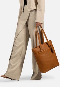Bolso tote de cuero marrón con doble asa, textura suave y un bolsillo lateral. Combinado con pantalones anchos beige y una chaqueta ligera.