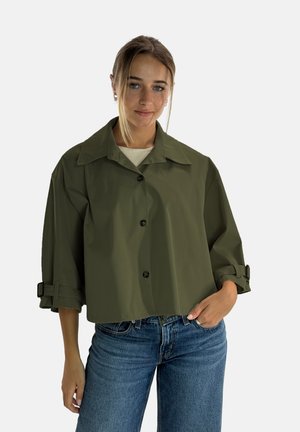Chaqueta corta de color verde oliva con cuello ancho, cierre de botones y mangas remangadas con correas ajustables. Combinada con vaqueros azules.
