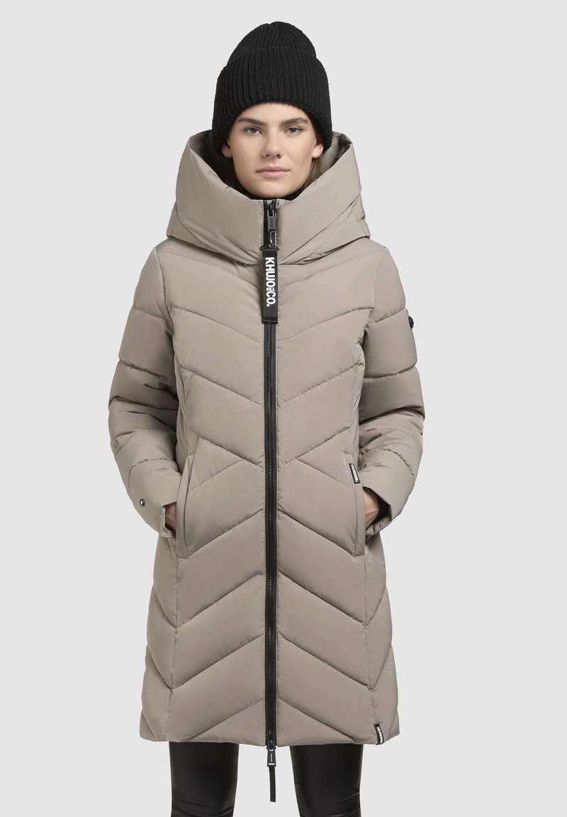 Padded coat in light beige with chevron pattern, large hood, and a black zipper. Features side pockets and a branded tag on the zipper pull.
