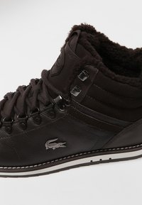 Bota de invierno de cuero marrón oscuro con forro de borreguito, ojales metálicos, suela gruesa y logo de cocodrilo en el costado.