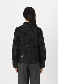 Casaco preto transparente com aplicações florais texturizadas e lantejoulas, gola com colarinho e mangas longas, combinado com calças cinzas às riscas.