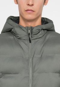 Αδιάβροχο μπουφάν σε απαλή πράσινη απόχρωση, με κλείσιμο μπροστά με φερμουάρ, θερμική επένδυση και κουκούλα. Λείος υφή με ορατές ραφές και λεπτομέρειες από φερμουάρ.