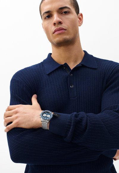 Tumesinine soonikkoes kampsun kraega kaelusega, mida täiendab hõbedase tooniga kell sinise sihverplaadi ja kronograafi kilpidega.