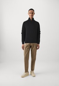 Schwarzer Half-Zip-Pullover mit geripptem Muster, kombiniert mit beigen Hosen und cremefarbenen Sneakers. Einfaches, modernes Design mit einer lockeren Passform.