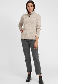 Strikkegenser i beige med høy krage, med to knapper. Kombinert med grå jeans og svarte ankelstøvler, som viser en avslappet look.