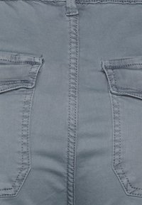 Grå denimjeans med flera fickor, synlig sömnad och en slät, strukturerad yta. Fokus på detaljerna bak och fickdesignen.