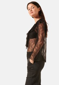 Blouse en dentelle noire avec des manches longues, un devant volé et un tissu transparent. Portée par-dessus un top noir, associée à un pantalon sombre.