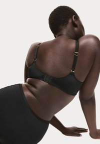 Sostenitore nero con spalline regolabili e dettagli in oro, con una texture liscia e un design della schiena a forma di U. Indossato con mutandine nere coordinate.