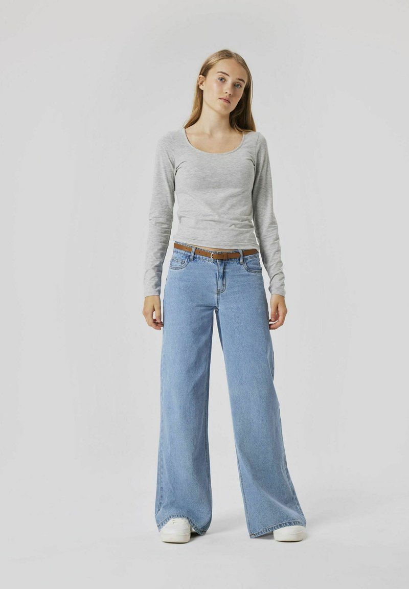 Haut gris à manches longues associé à un jean large bleu clair, comportant une ceinture marron et des baskets blanches, sur fond neutre.