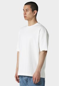 Hvit bomull T-skjorte med avslappet passform, korte ermer og rund hals. Har en liten etikett på ermet, kombinert med blå jeans.