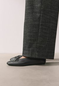 Bailarinas de cuero negro con punta afilada y detalle decorativo de lazo. Usadas con pantalones de pierna ancha con estampado gris sobre una superficie neutra.