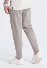 Pantalones de chándal gris claro con puños elásticos, bolsillos laterales y textura suave, combinados con zapatillas blancas, vistos desde la parte posterior.