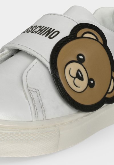 Бял спортен обувка с черно лого Moschino на каишката и кафяв нашивка с анимационен мечок на страната.