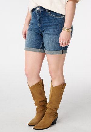 Vrouw draagt opgerolde denim shorts gecombineerd met beige suède halfhoge laarzen, gouden armbanden en meerdere ringen, staand tegen een witte achtergrond.