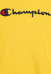 Felpa gialla con cappuccio presenta una tasca a canguro frontale e un logo "Champion" ricamato in contrasto, in blu navy con un accento rosso.