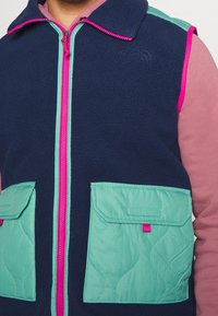 Marineblaue Fleece-Weste mit tealfarbener Schulterpartie und Taschen, ausgestattet mit einem pinkfarbenen Reißverschluss und Akzenten. Das Material hat eine weiche Textur und ein gestepptes Taschendesign.