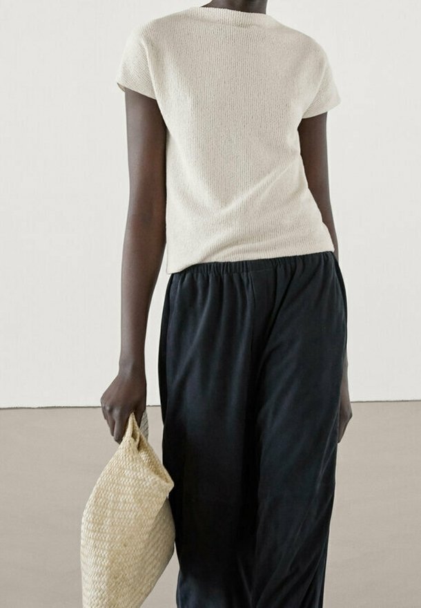Personne portant un haut blanc texturé à manches courtes et un pantalon noir ample, tenant un grand sac cabas beige tissé à côté d'elle.