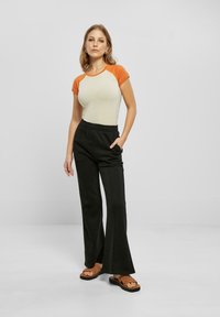 Lys beige topp med oransje ermer, svarte vide bukser og brune sandaler med utskjæringsdesign, stående mot en ensfarget bakgrunn.