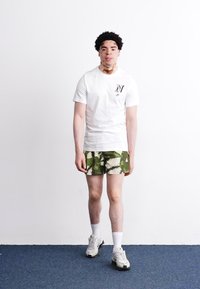 Hvid kortærmet t-shirt med logo på brystet, kombineret med grønne camouflage shorts. Bærer hvide sokker og klodsede hvide sneakers.