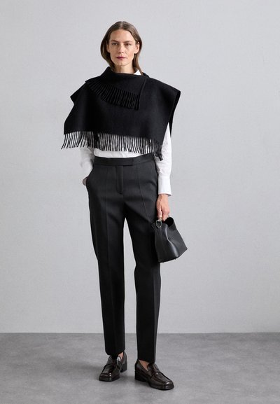 Black wool poncho with fringed hem, worn over a white shirt. Paired with tailored black trousers and a small black leather bag. Brown shoes.