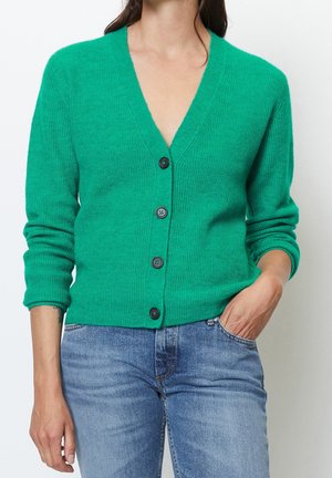 Cardigan vert côtelé avec un col en V, des manches longues et des boutons noirs. Porté par-dessus un jean bleu, avec une coupe décontractée et une texture douce.