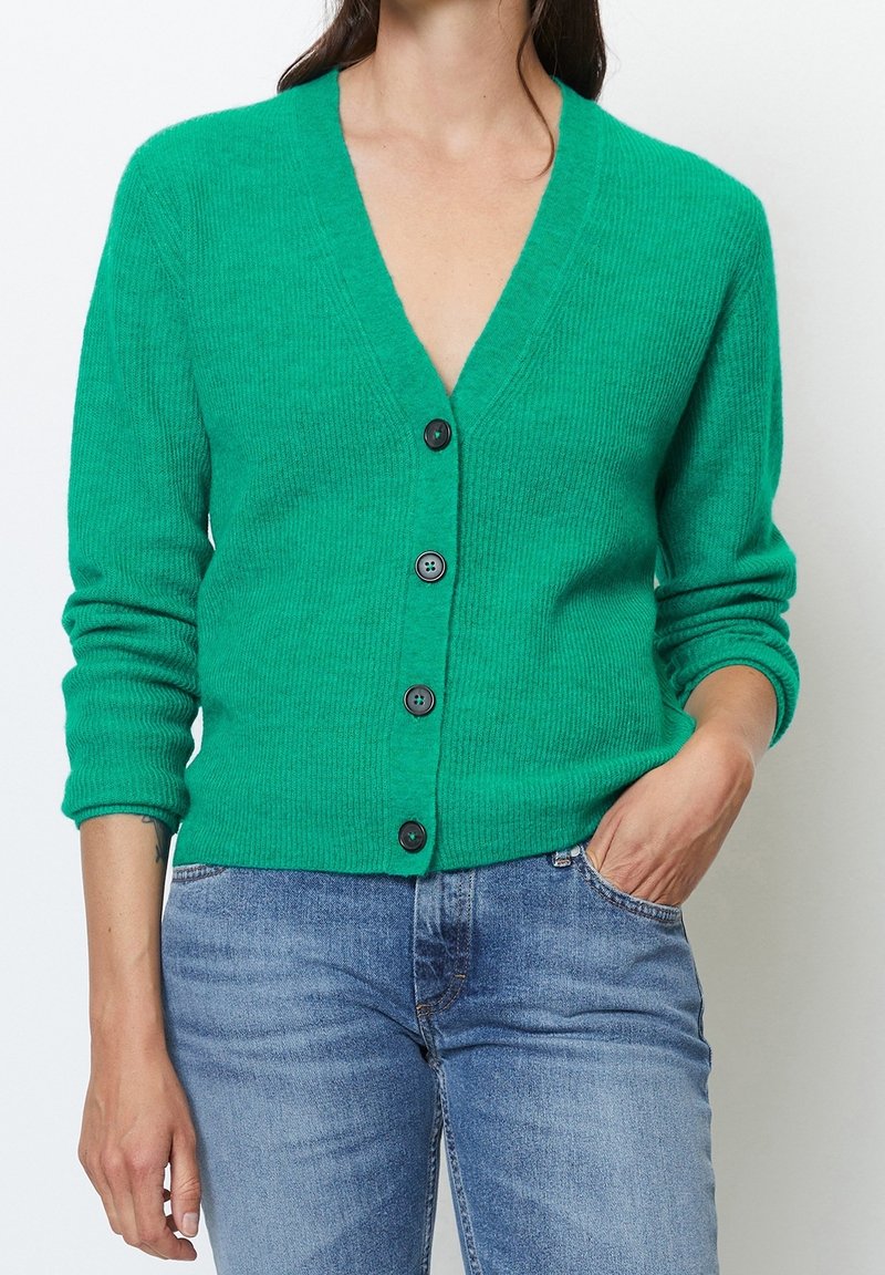 Cárdigan verde con canalé, de cuello en V, mangas largas y botones negros. Llevado sobre jeans azules, con un ajuste relajado y una textura suave.