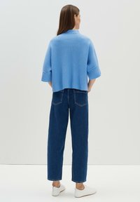 Blauer gerippter, verkürzter Pullover mit weiten Ärmeln, kombiniert mit hoch taillierten, dunklen Jeans und weißen Sneakern. Einfarbiger Hintergrund.