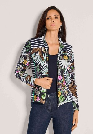 Vrouw die een bloemen- en gestreepte ritsjack draagt over een zwarte top, gecombineerd met donkerblauwe spijkerbroek, staand tegen een effen achtergrond.