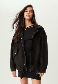 Une jeune femme aux longs cheveux noirs porte une veste noire surdimensionnée par-dessus une robe noire avec des œillets en métal au niveau de l'encolure.