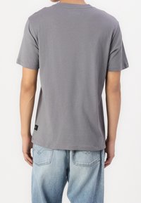 Grijze katoenen t-shirt met korte mouwen, ronde hals en een rechte snit. Opmerkelijke label-details aan de onderzoom, gedragen met lichtblauwe jeans.