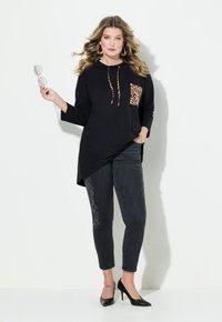 Túnica negra con capucha y un bolsillo de estampado de leopardo, combinada con jeans oscuros con acentos decorativos. Modelo con tacones negros, sosteniendo gafas de sol.