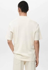 Lyhythihainen, kermaisenvärinen t-paita, valmistettu pehmeästä kankaasta, jossa rento istuvuus ja yksinkertainen pyöreä kaula-aukko. Takaisinäkymä, jossa on minimaalinen yksityiskohtaisuus.